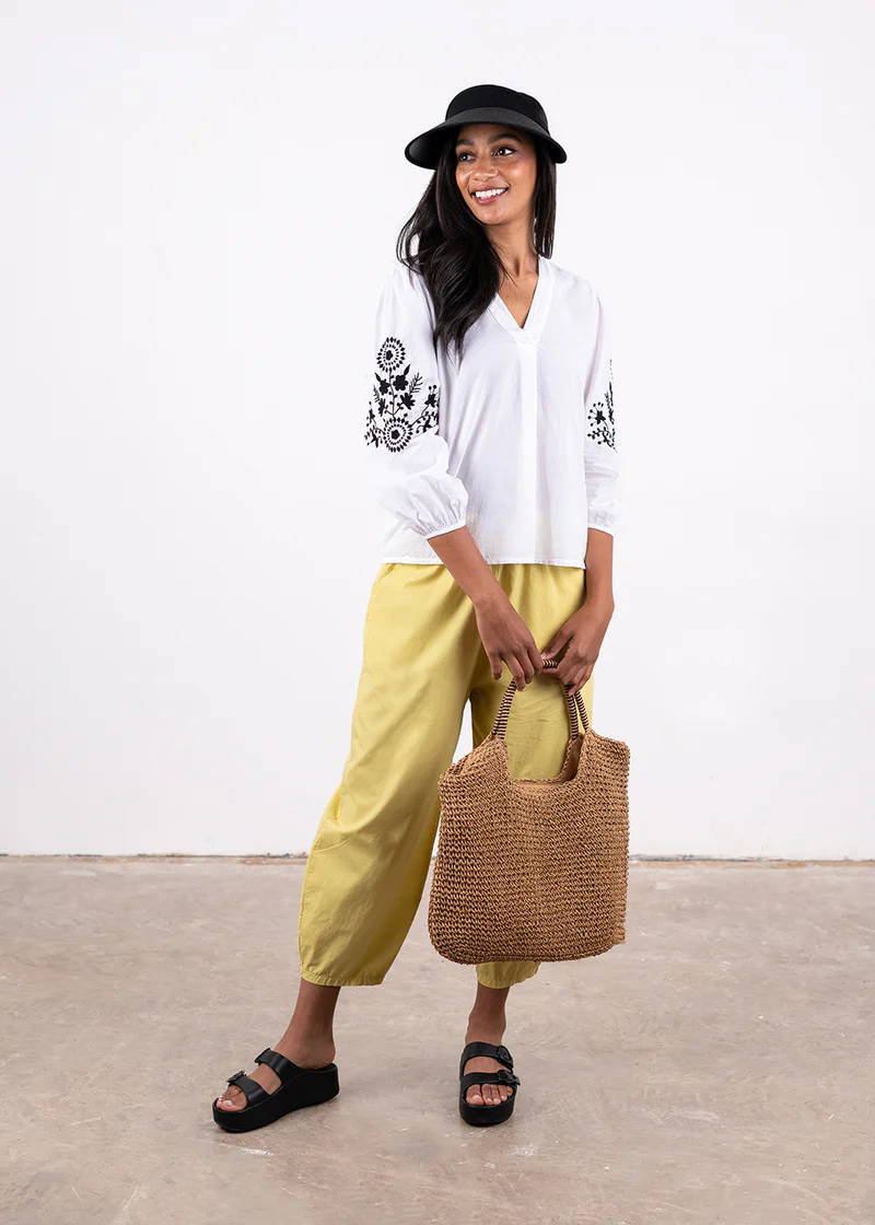 A model wearing a white long sleeved blouse with a v neck and black embroidered detailing on the outer arm, pale yellow barrel leg trousers and black chunky platform slides