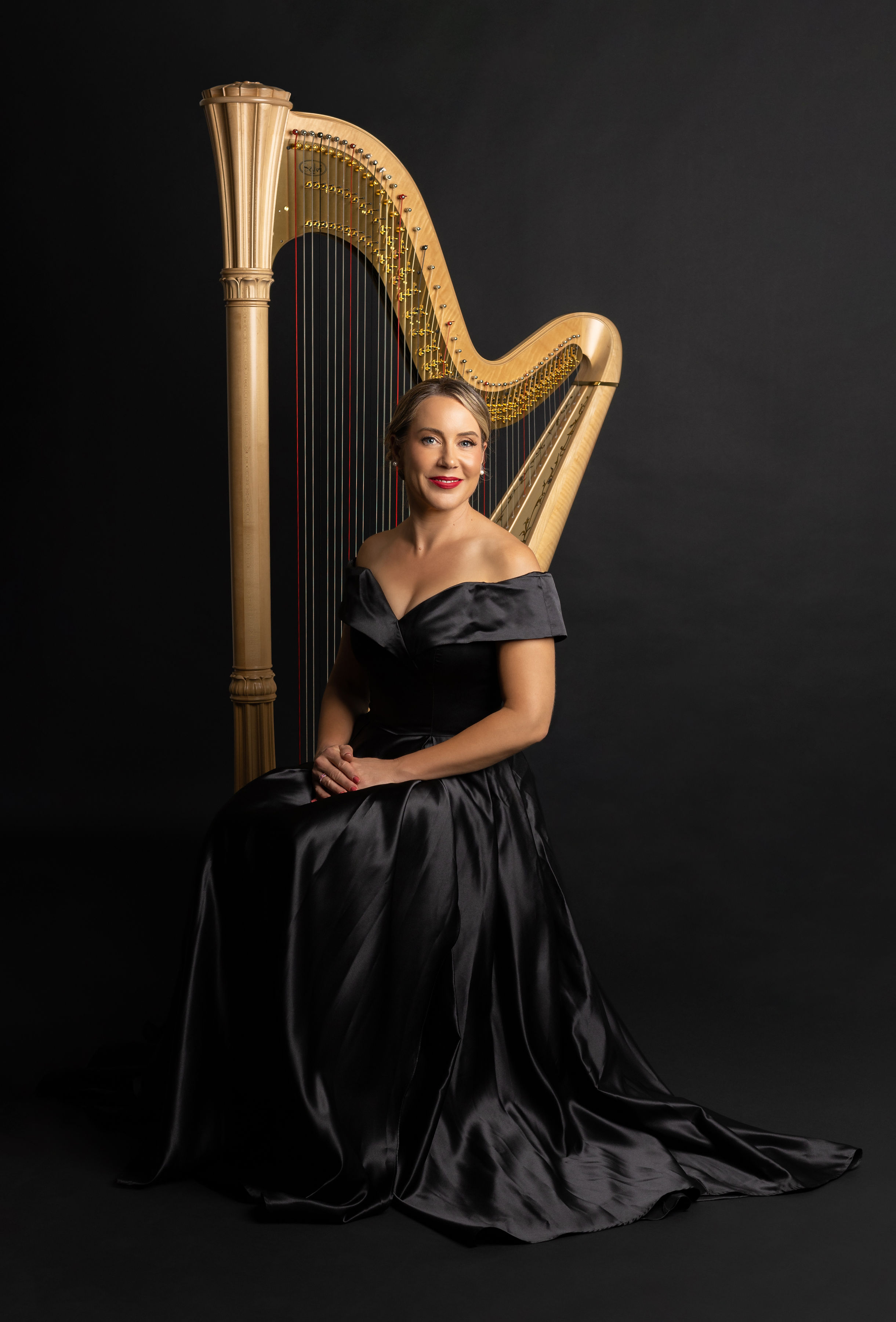 Woman in black gown sitting beside a harp against a dark background.