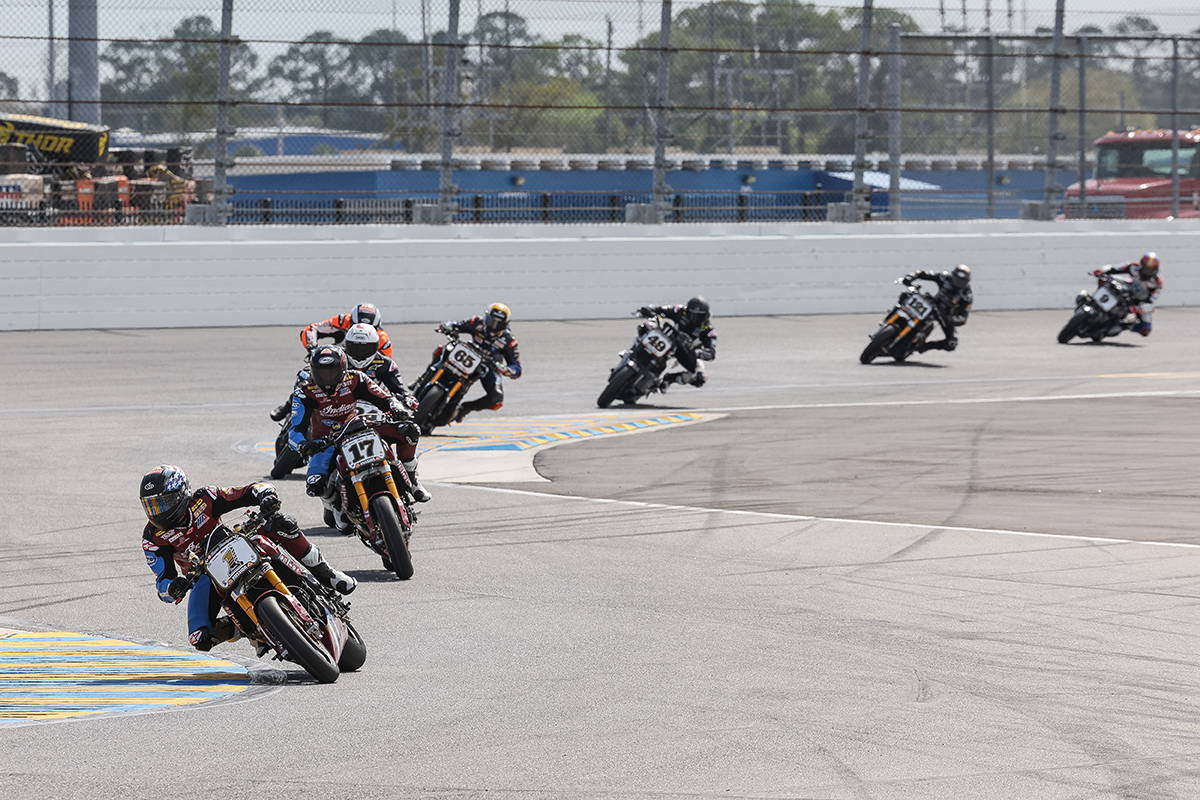 Aerial view of a motorcycle racer clad in a maroon and gold suit, crossing the Daytona International Speedway's finish line marked by a checkerboard pattern on the ground.