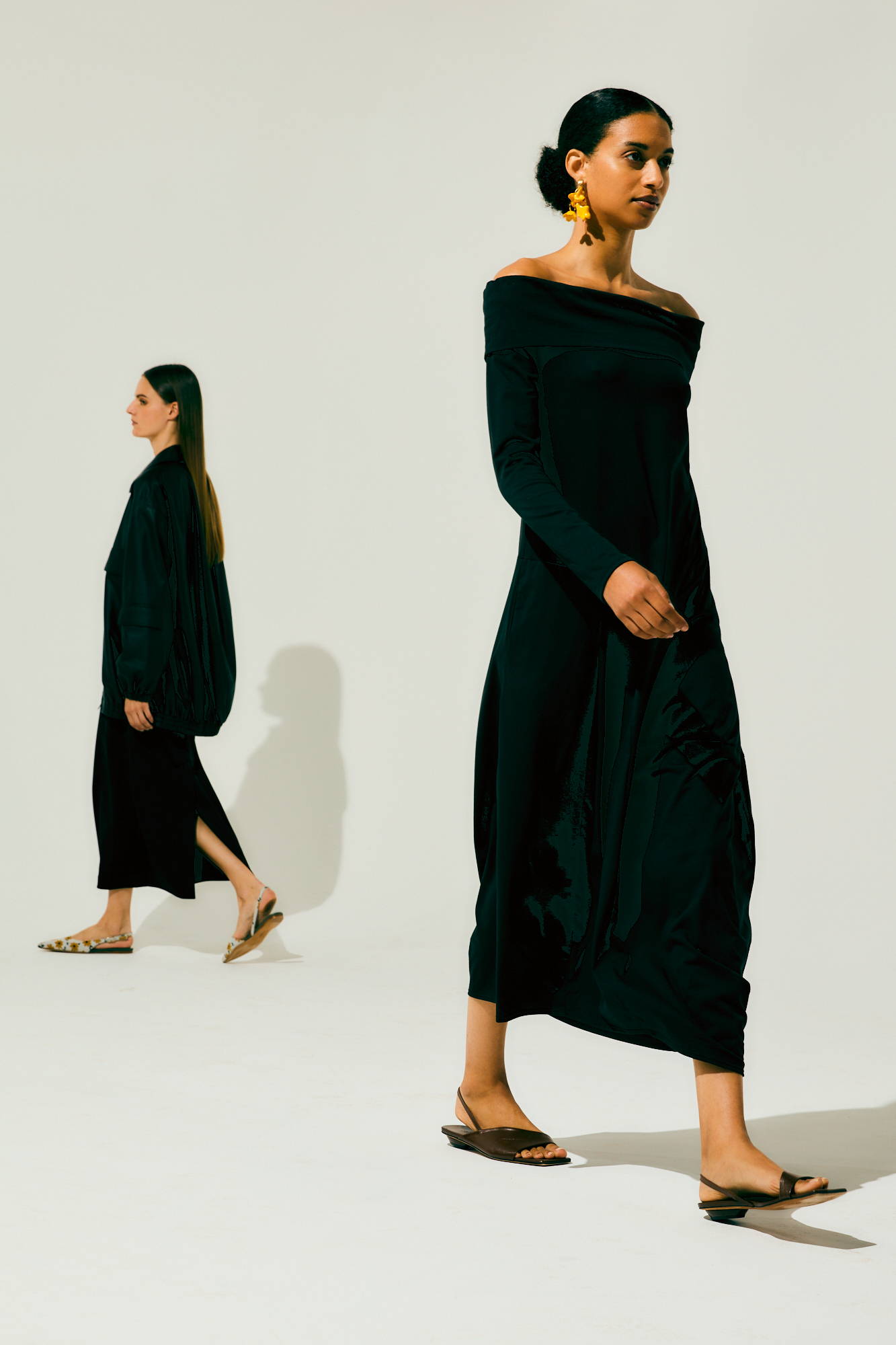 Two models walking in black dresses against a neutral background.