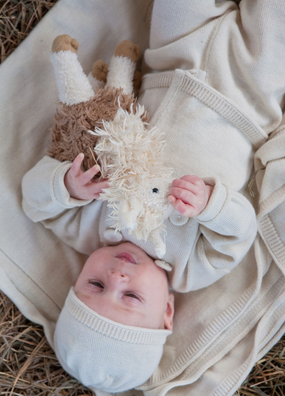 Baby lying on a blanket, holding a stuffed animal, surrounded by hay.