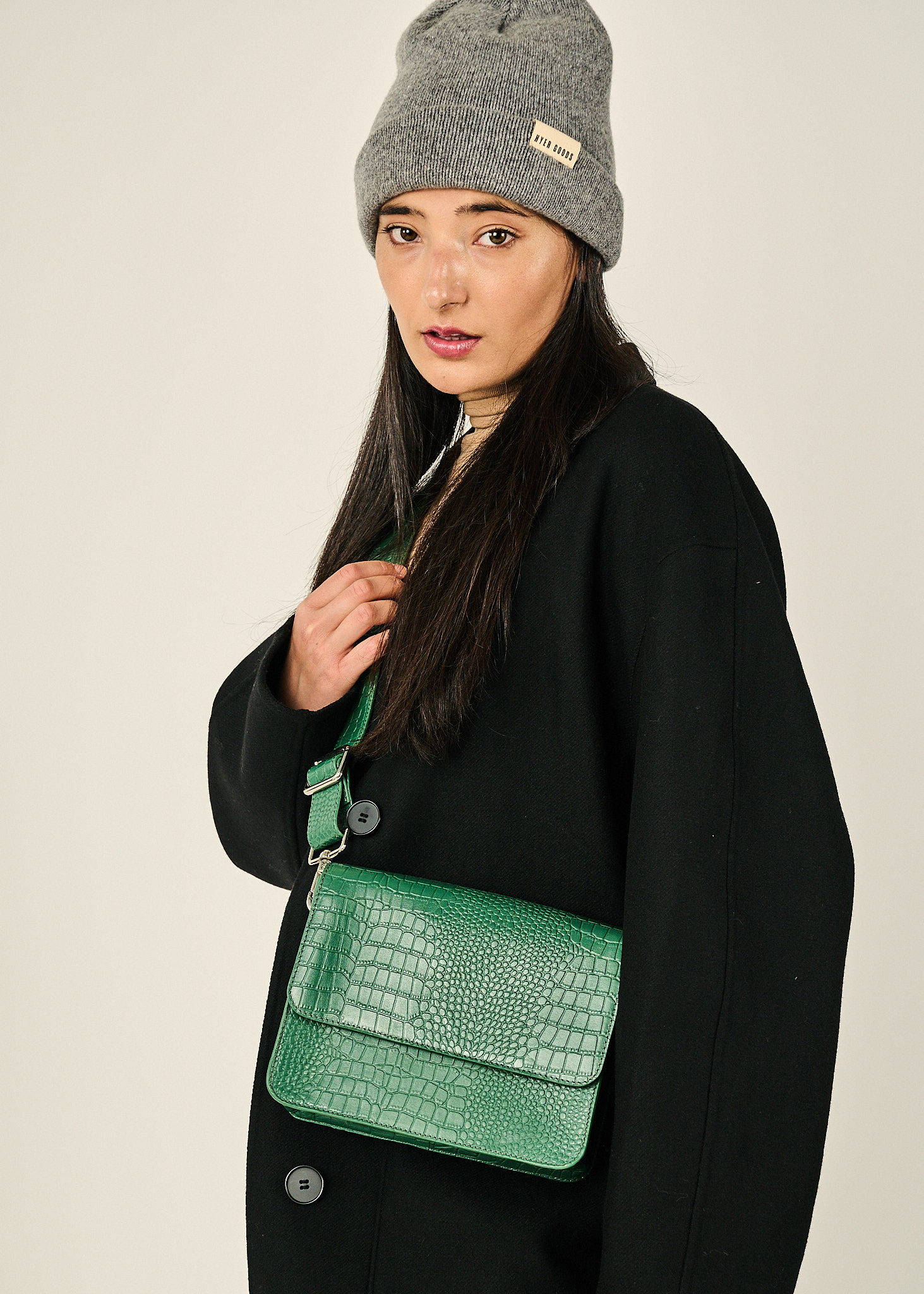 a 3/4 view of a girl wearing a grey wool beanie, a black coat and a bright green crocodile crossbody purse