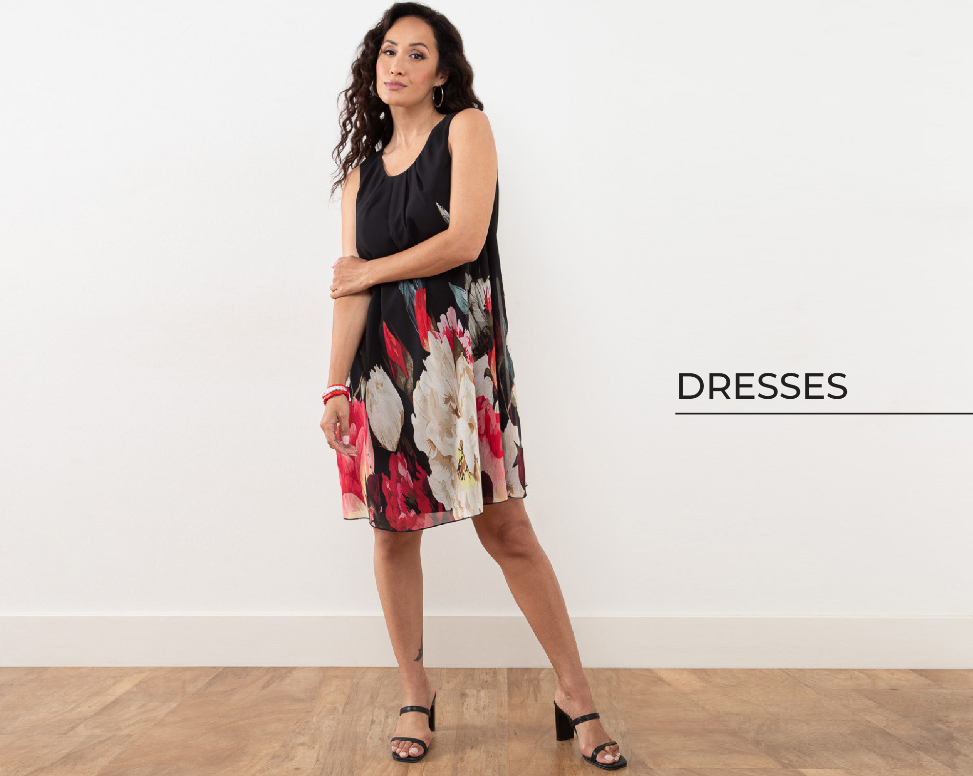 Woman wearing a floral dress, standing on wooden floor with a white background.