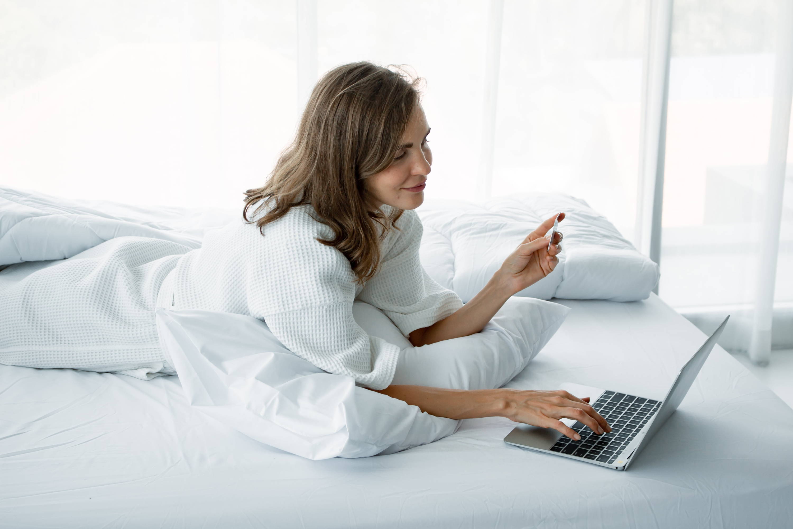 Woman lies on a pillow on her front in bed. One hand is typing on a laptop and the other holds a credit card.