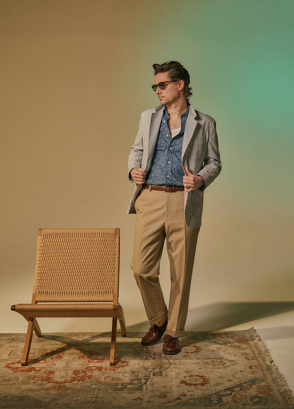 A man in a blazer and sunglasses poses beside a chair.