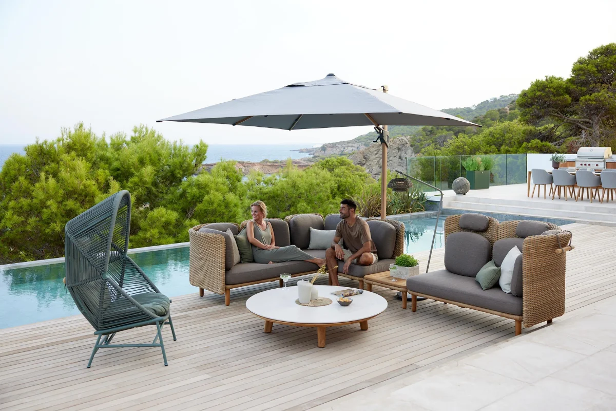 Resort-style poolside lounge with woven outdoor furniture and large cantilever umbrella providing shade over a luxury patio overlooking coastal-inspired landscaping.