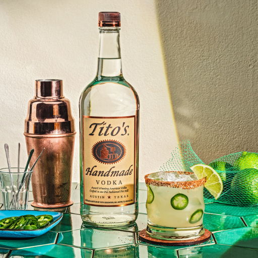 A bottle of Tito's vodka with a cocktail and garnishes on a table.