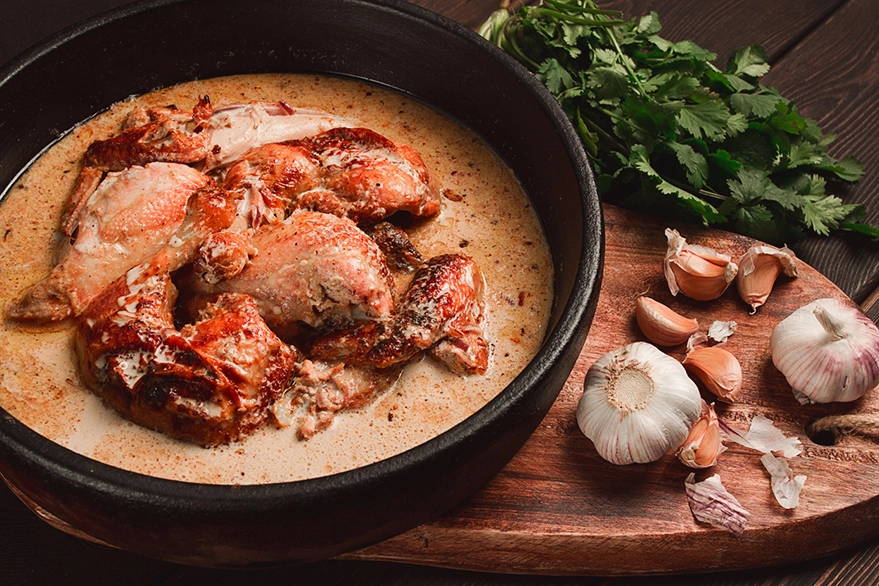 Traditional Georgian Shkmeruli, roasted chicken in a garlic milk sauce, accompanied by garlic cloves and parsley on a wooden backdrop, showcasing the rich culinary traditions of Georgia.