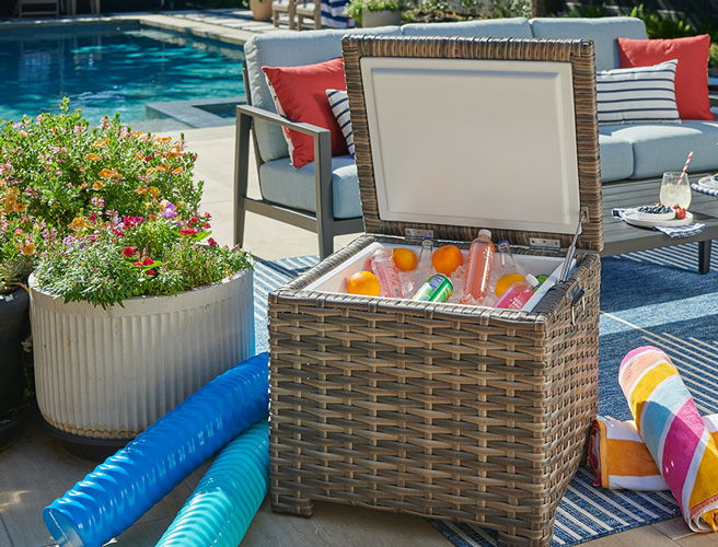 Woven cooler by poolside with drinks and colorful pool floats nearby.