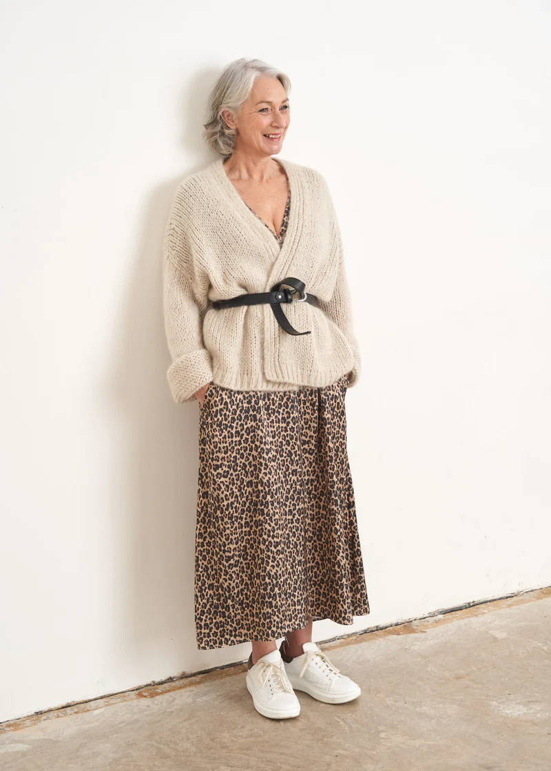 A model wearing an off white knitted slouchy cardigan secured with a black belt over a leopard print midid dress and white trainers