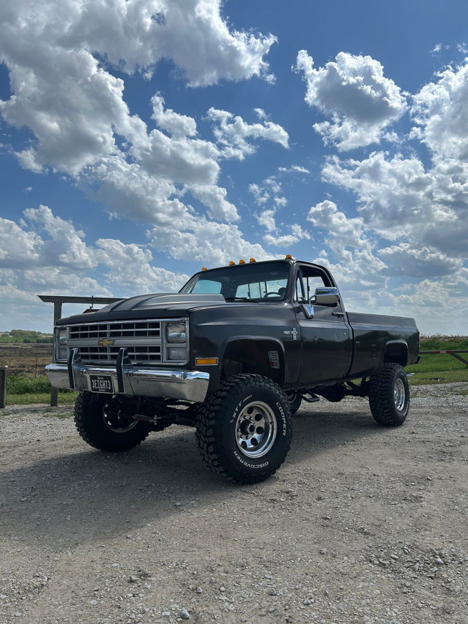 Low-angle three-quarter front view Kyle's black 1985 Chevy K20 Deluxe Pickup lifted with raised cowl hood chrome grille and Cooper Discoverer off-road tires on rural gravel HornBlasters Conductor's Special 127H featured customer install gallery photo 1