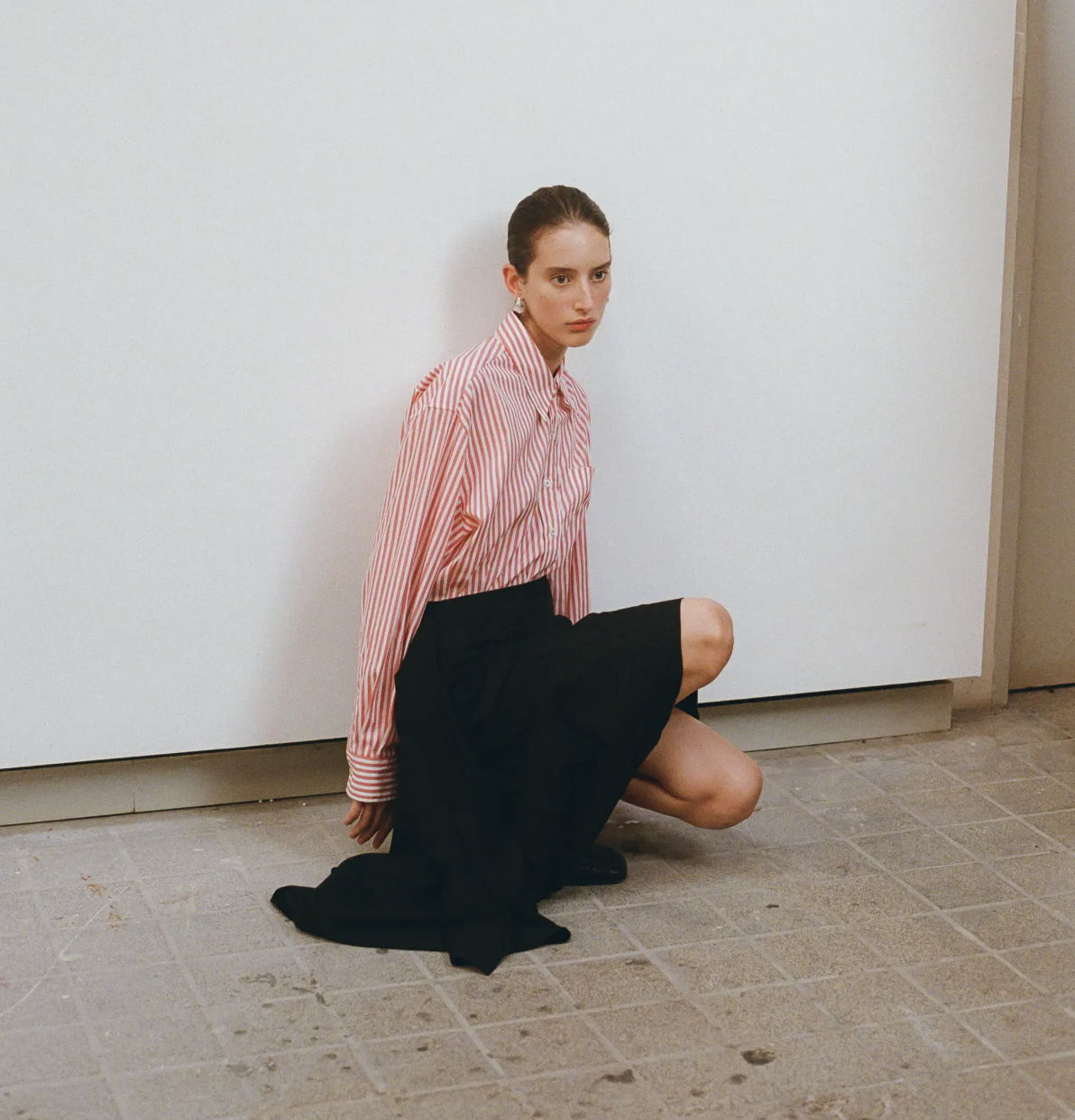 A model in a pink shirt and black skirt poses against a white wall.