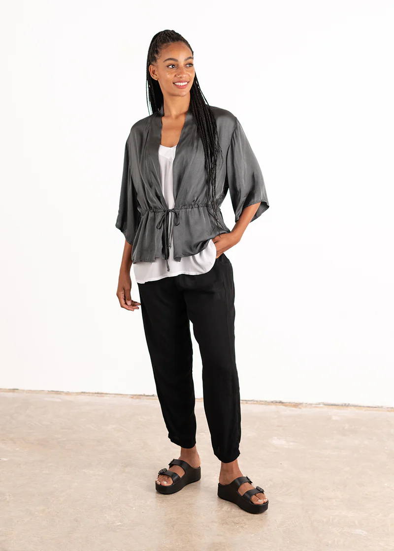 A model wearing a dark grey satin, loose fitting jacket with a draw string waits and 3/4 length sleeves over a white top, black trousers and black slides