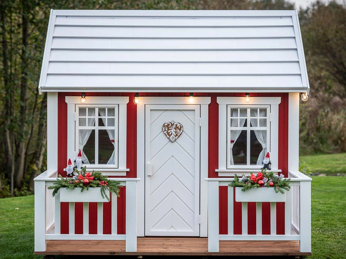 Red and white wooden playhouse with a white porch and Christmas decorations with forest background by WholeWoodPlayhouses