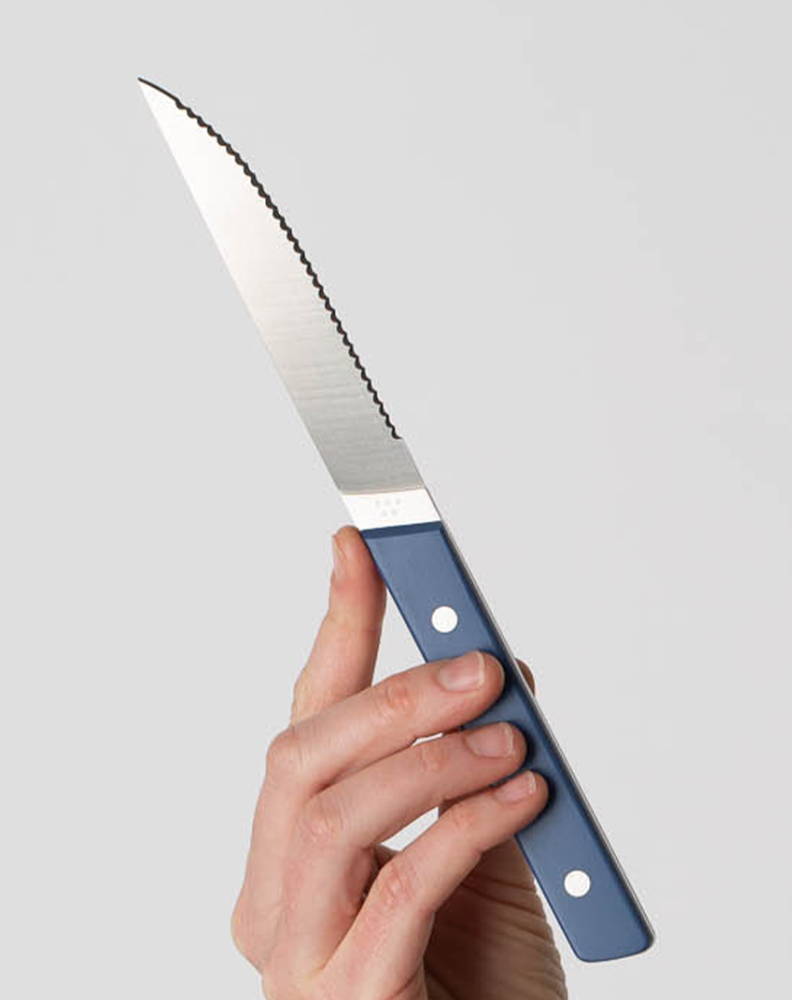 A hand holding a Blue Misen Steak Knife vertically with the blade’s tip facing upwards.