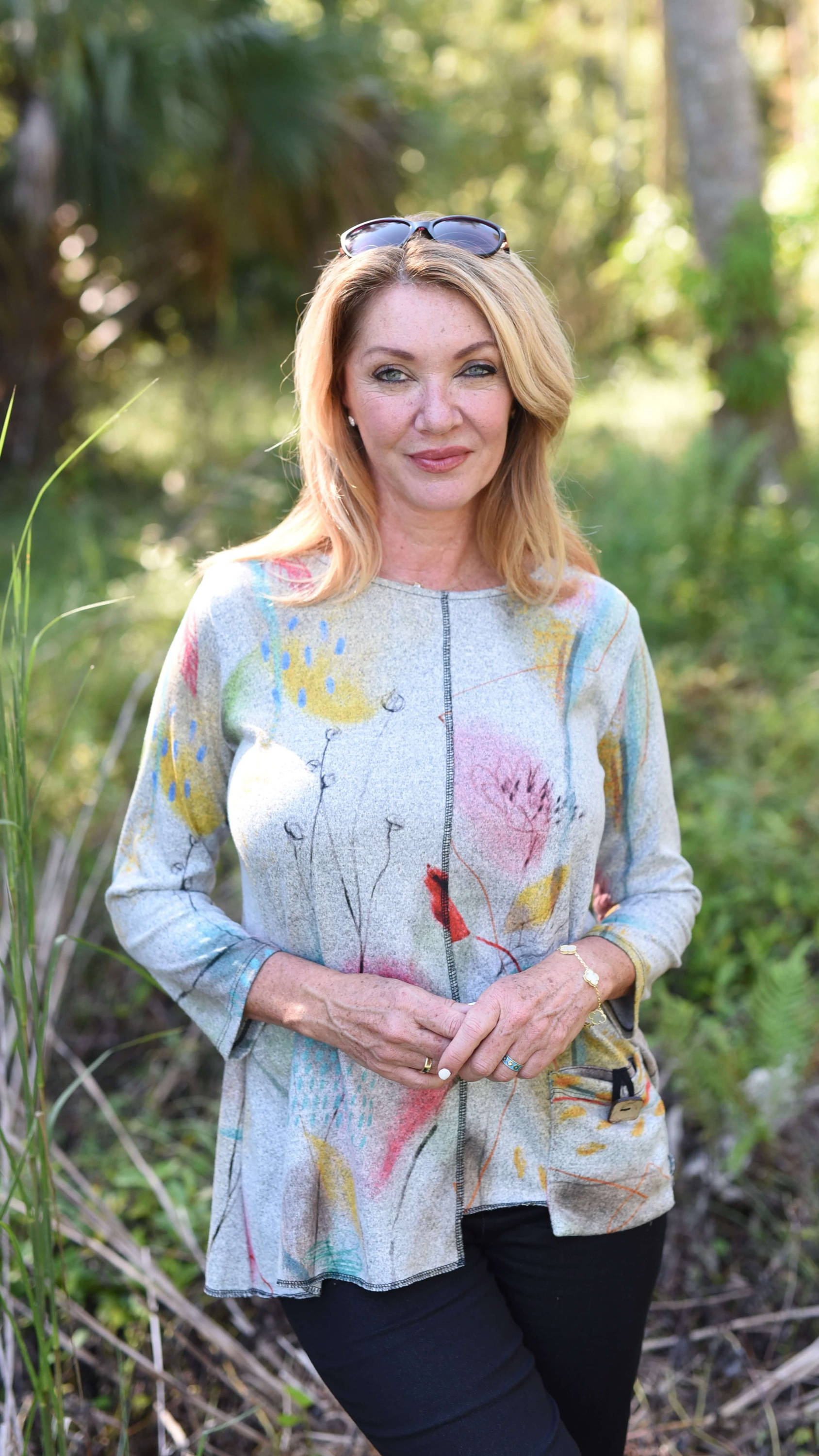 A blonde woman with sunglasses perched on the top of her head stands in a forested area and clasps her hands together. 