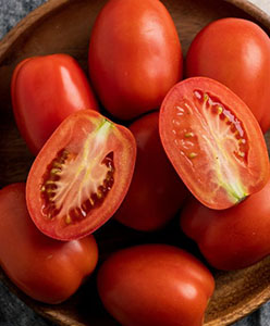 Juicy red tomatoes arranged in a wooden bowl, some halved revealing seeds.