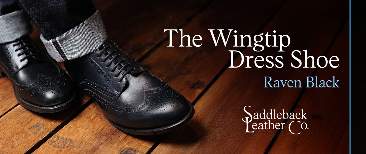 Black wingtip dress shoes on a wooden floor, featuring rolled-up pants.