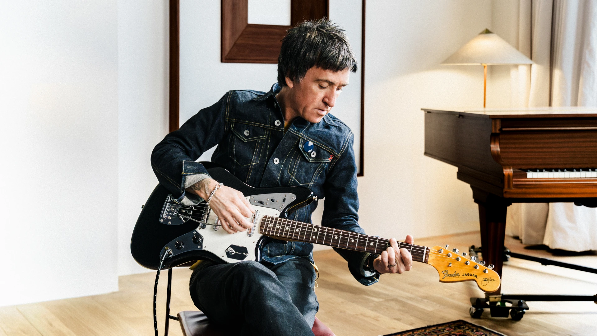 Johnny Marr playing his Fender signature Jaguar electric guitar in a room with a piano.