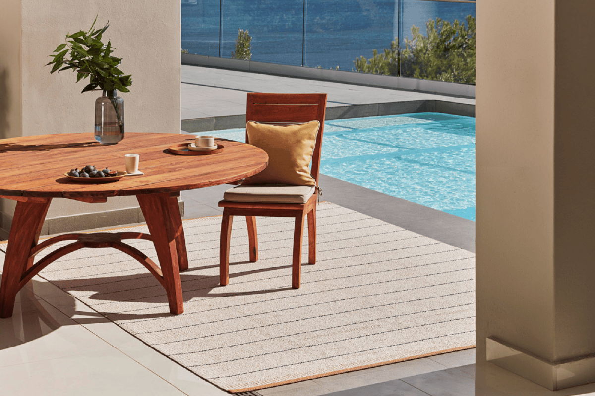 Neutral striped outdoor rug under a round wood dining table and chair on a covered patio beside a pool, showing how an outdoor rug grounds a dining area.