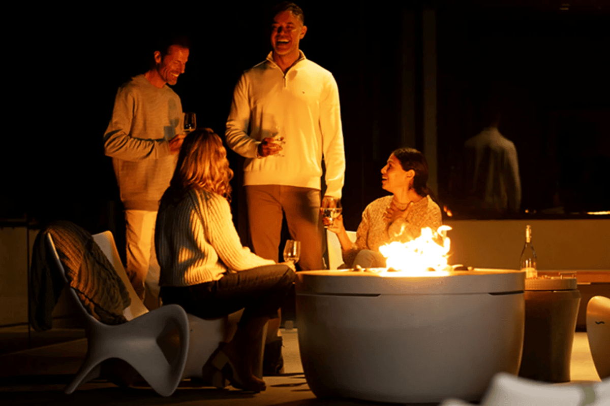 Guests gathered around a modern outdoor fire pit at night, extending a summer backyard gathering with warm firelight.
