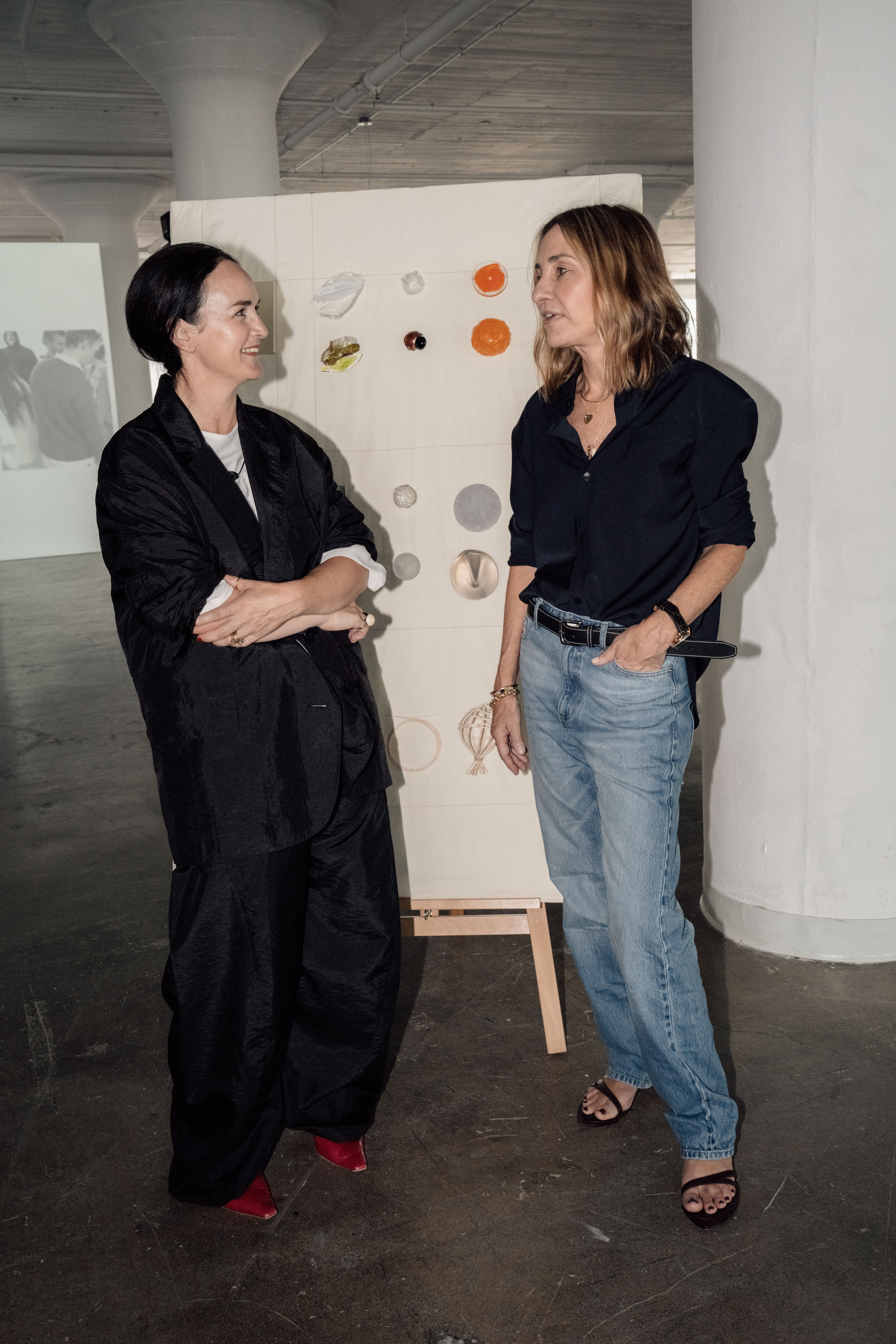 Two women engaged in conversation near an art display in a gallery.