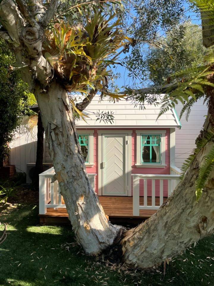 Pink Wooden Playhouse Unicorn with white metal roof and wooden terrace with railing on green lawn in a backyard by WholeWoodPlayhouses 