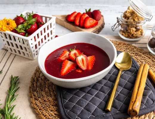 Bowl of red soup topped with strawberries, surrounded by fresh strawberries and granola.