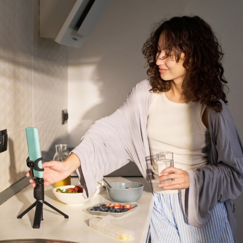 Woman holding a glass of water while creating content with a smartphone in her kitchen.