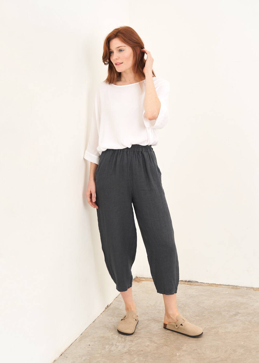 A model wearing a white long sleeve top with dark grey cropped linen trousers and taupe coloured clogs