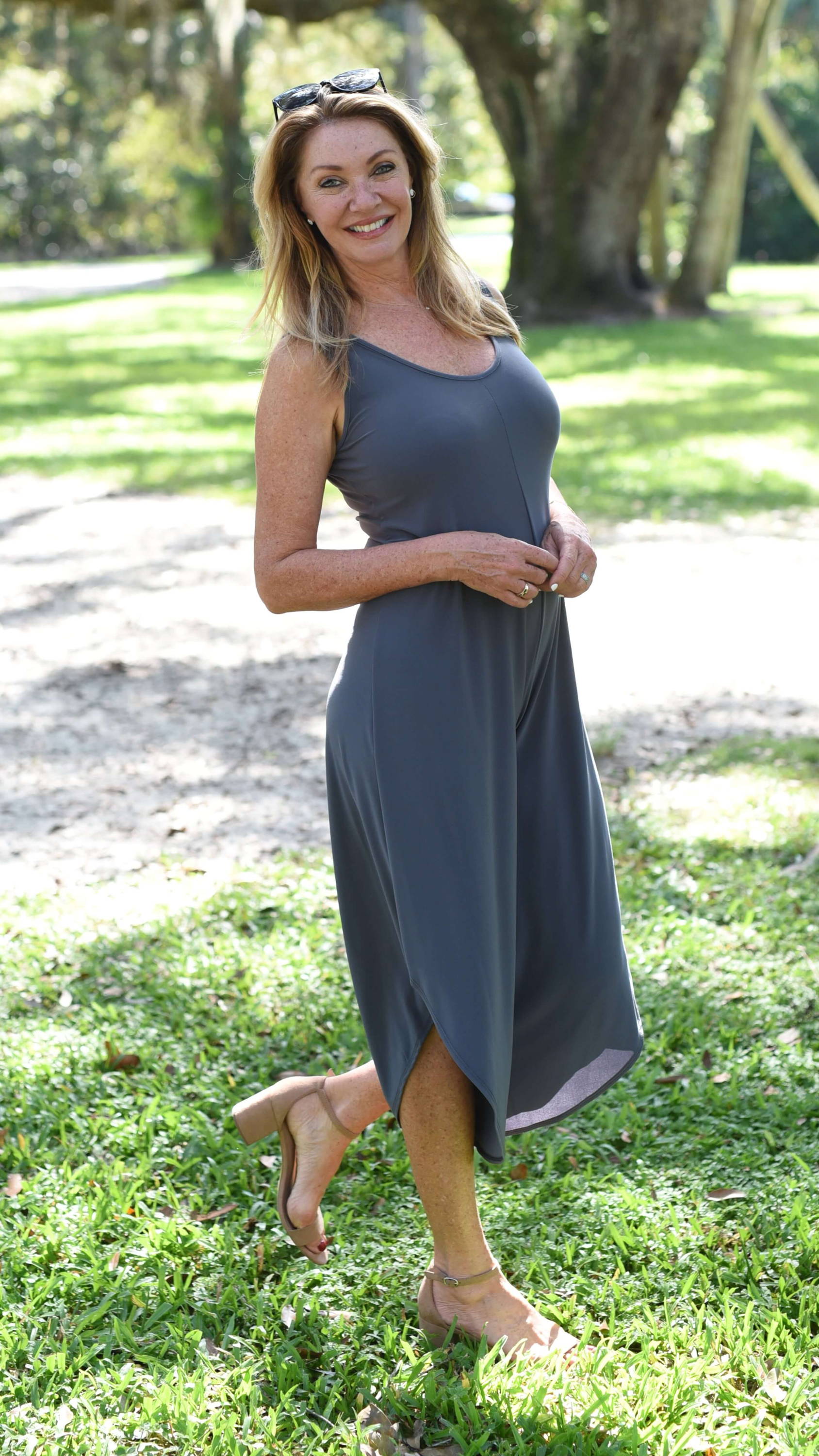 A smiling blonde woman in a grey jumpsuit sands in a forested area. 