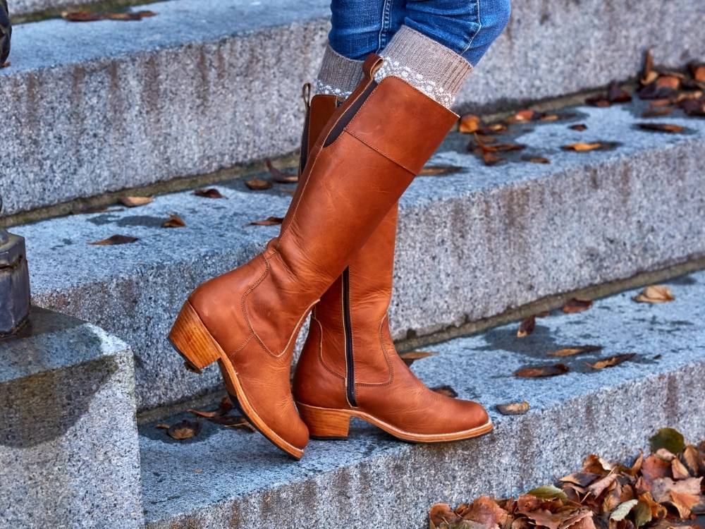 An outdoor autumn lifestyle product shot from the waist down of a person standing on wet stone stairs scattered with fallen leaves. The person is wearing blue jeans and thick knit grey boot socks peeking out from the top of tall, knee-high tan leather boots featuring a stacked block heel, a rear pull tab, and a dark side zipper.