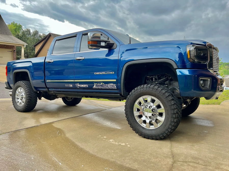 Front-three-quarter low-angle Kolyn's metallic blue lifted 2016 GMC Sierra Denali HD crew cab wet driveway chrome wheels dark headlights DENALI HD HornBlasters R1 Concepts door decals brick house trees cloudy sky Conductor's Special 244K Spare Tire Delete Kit photo 1