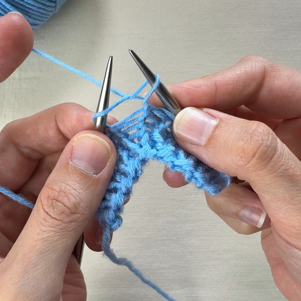 Detailfoto, wie eine rechte Masche gestrickt wird. Es hat sich eine rechte Masche gebildet, die nun auf der rechten Nadel liegt.