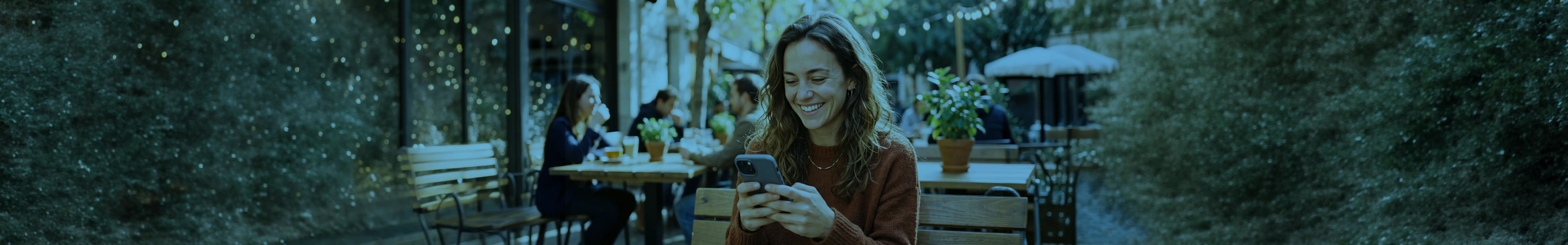 A woman smiling while holding a phone in a natural setting.