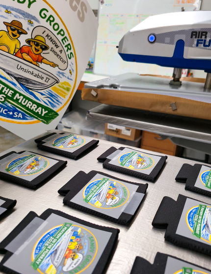 A spread of stubby coolers with cut logos printed on DTF film layed out ready for heatpressing