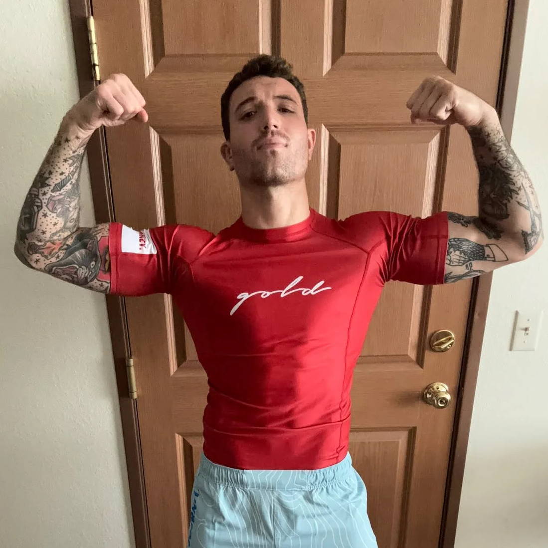 Man flexing muscles in a red shirt and shorts, standing in front of a door.