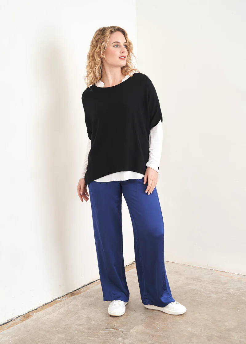A model wearing a pair of blue satin long wide leg trousers with a black sleeveless sweater over a white long sleeved top and white trainers