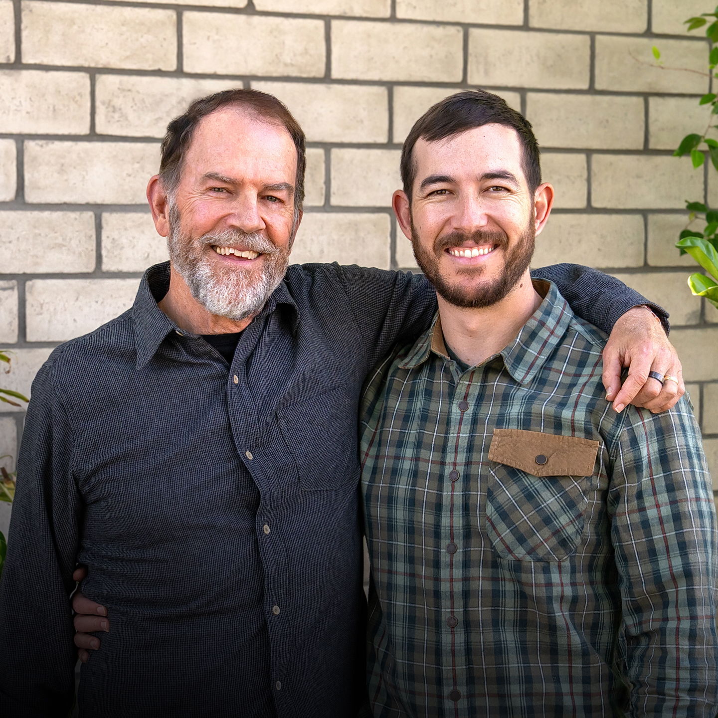 Two men smiling together in front of a stone wall.
