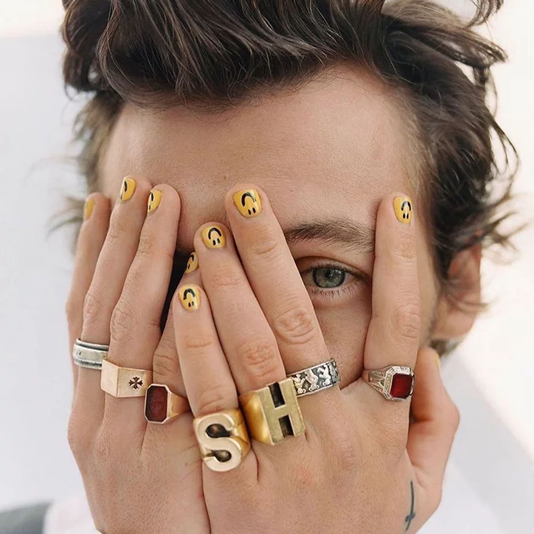 Person covering face with hands adorned in rings and yellow nail polish.