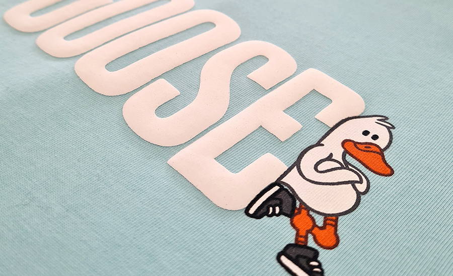 A close up of specialty puff ink screen print of the word GOOSE in white on a light blue shirt. There is a white cartoon duck with his arms crossed and an orange beak and legs, wearing black high top sneakers leaning against the raised texture letter E
