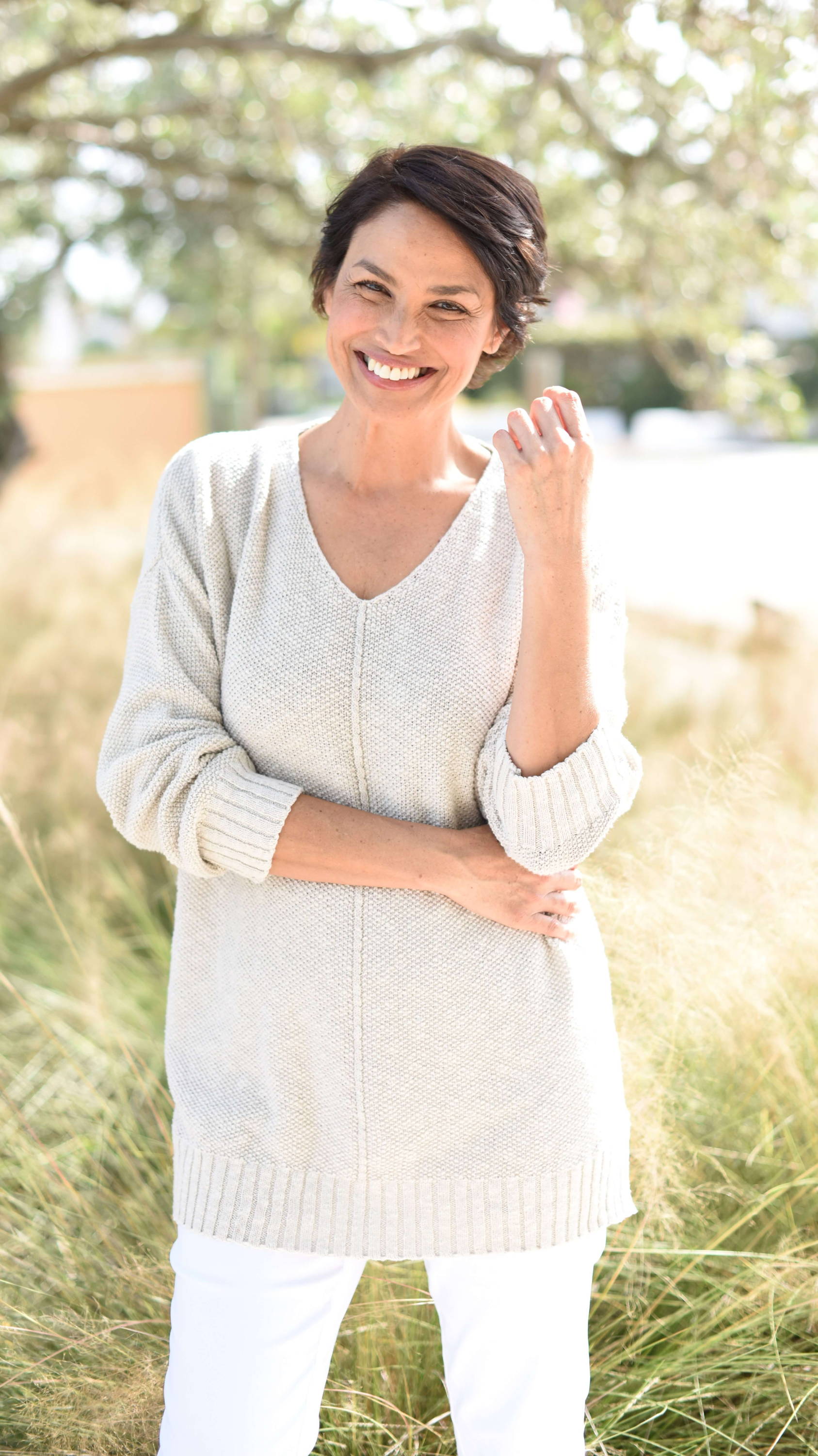 A smiling woman with short dark hair wears a tan sweater and white pants as she stands in a field