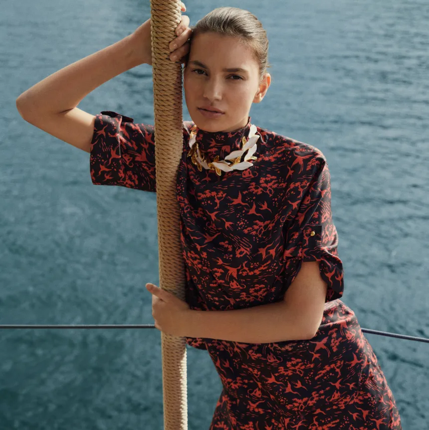 Model posing by the water, wearing a patterned dress and necklace.