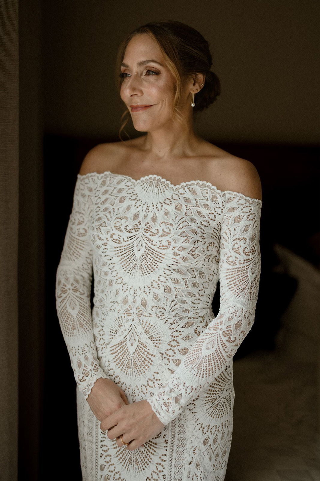 Woman in an elegant off-shoulder lace wedding dress, standing by a window.