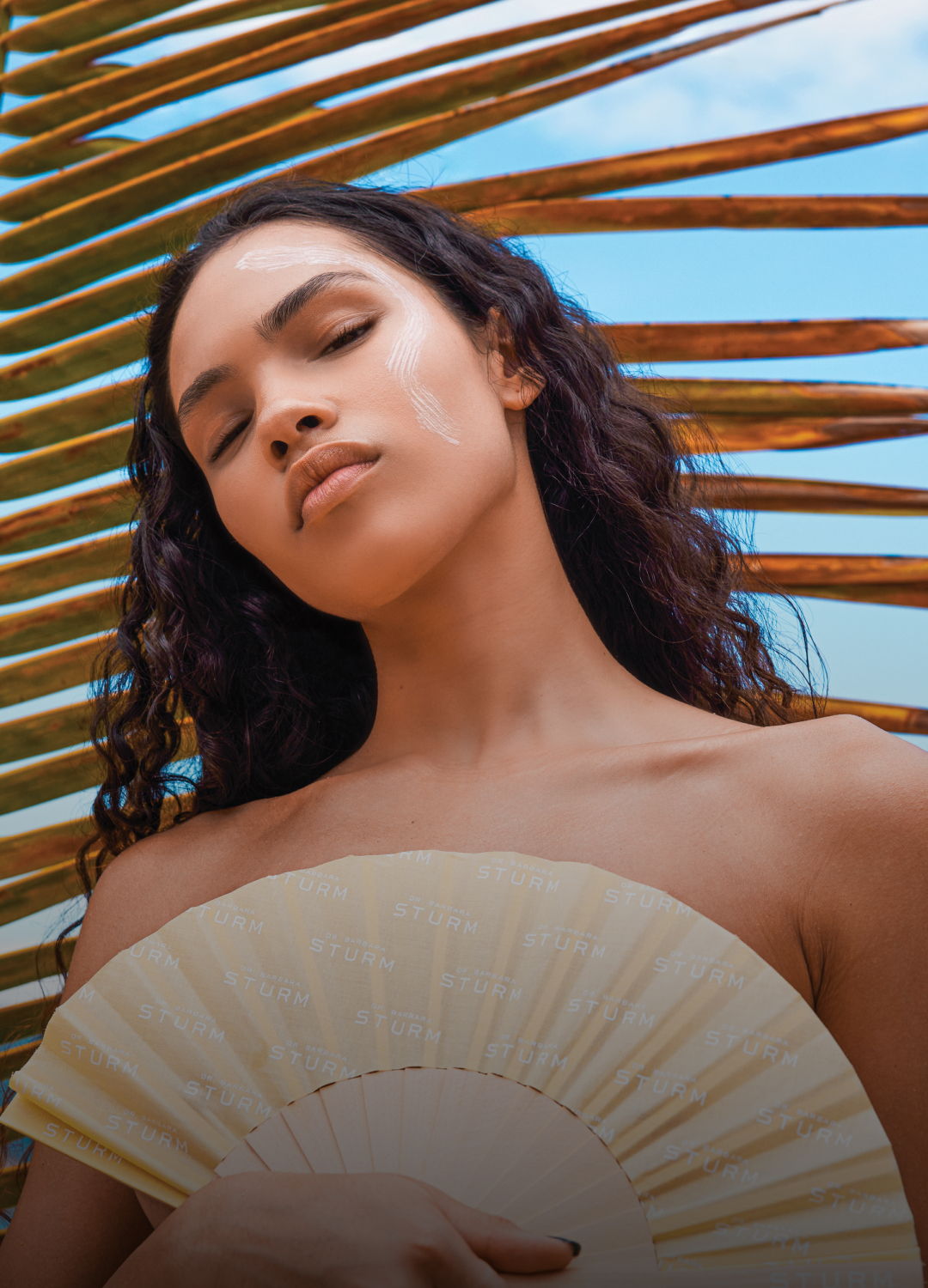 Woman with curly hair holding a fan, surrounded by palm leaves and blue sky.