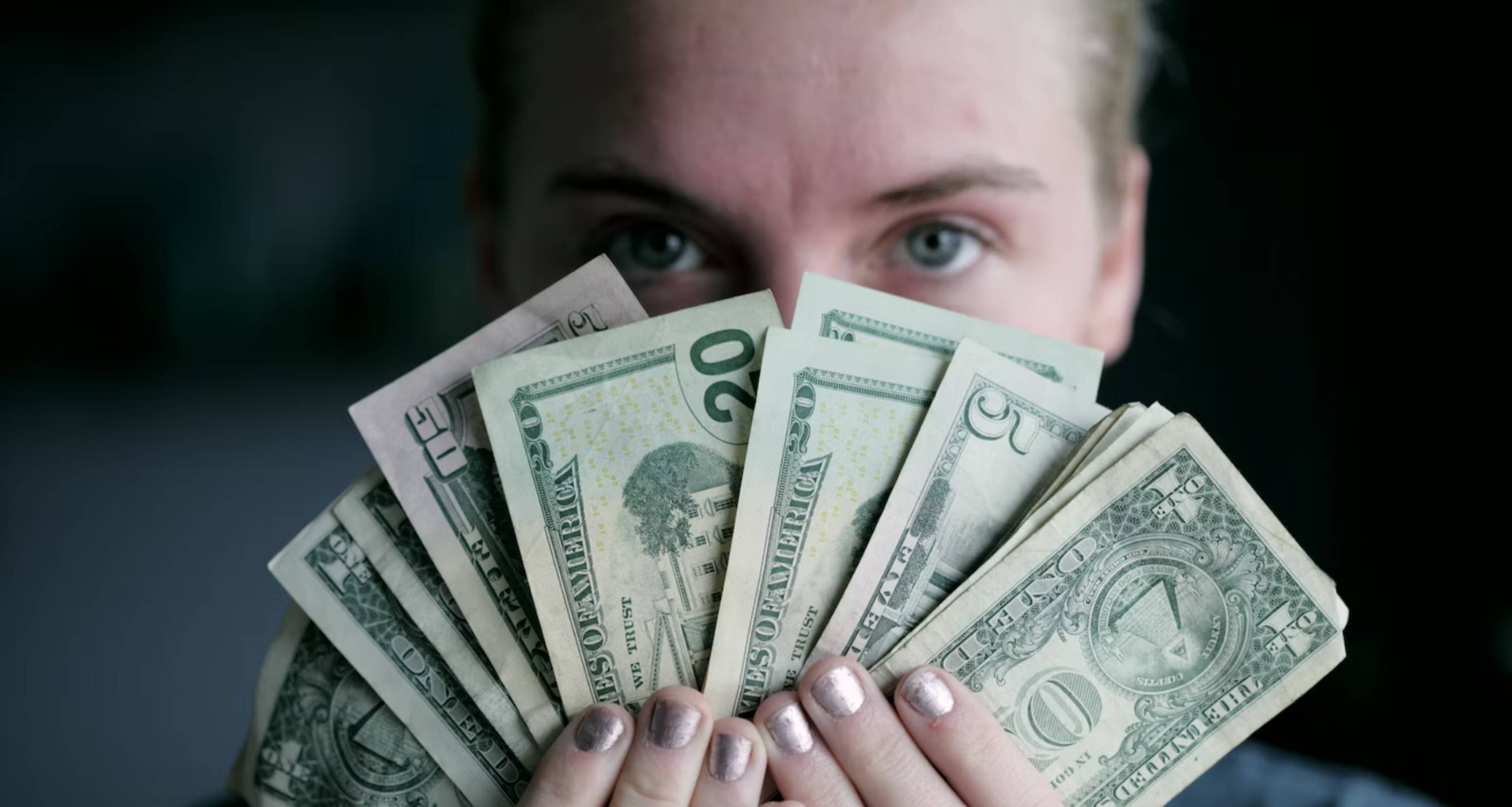 : blue-eyed woman holds a fan of dollar bills in front of her face