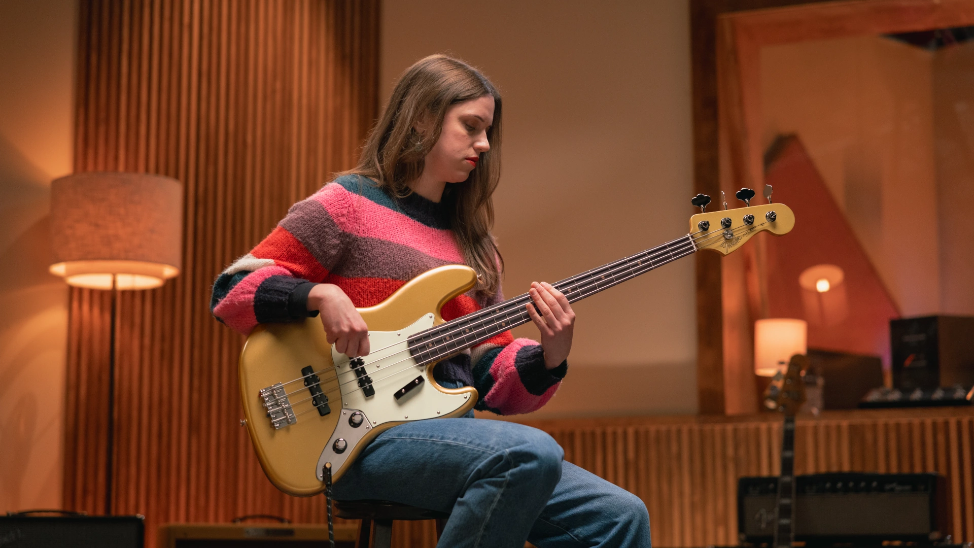 A woman playing a gold Fender Vintera III Early '60s Jazz Bass bass guitar in a studio setting.