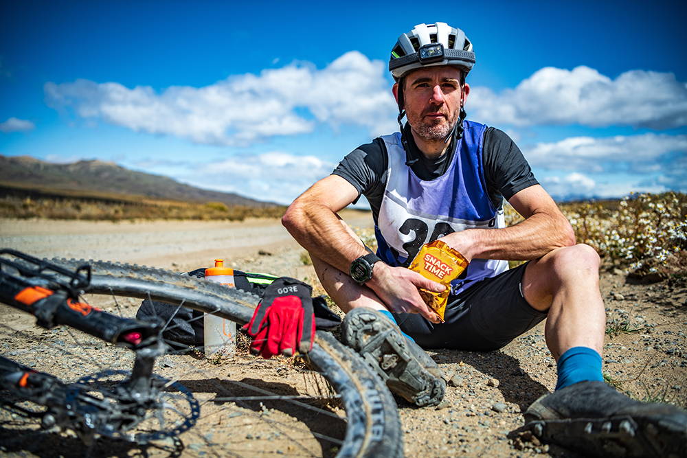 A cyclist sitting next to a gravel road next to his bike with helmet on and eating a snack
