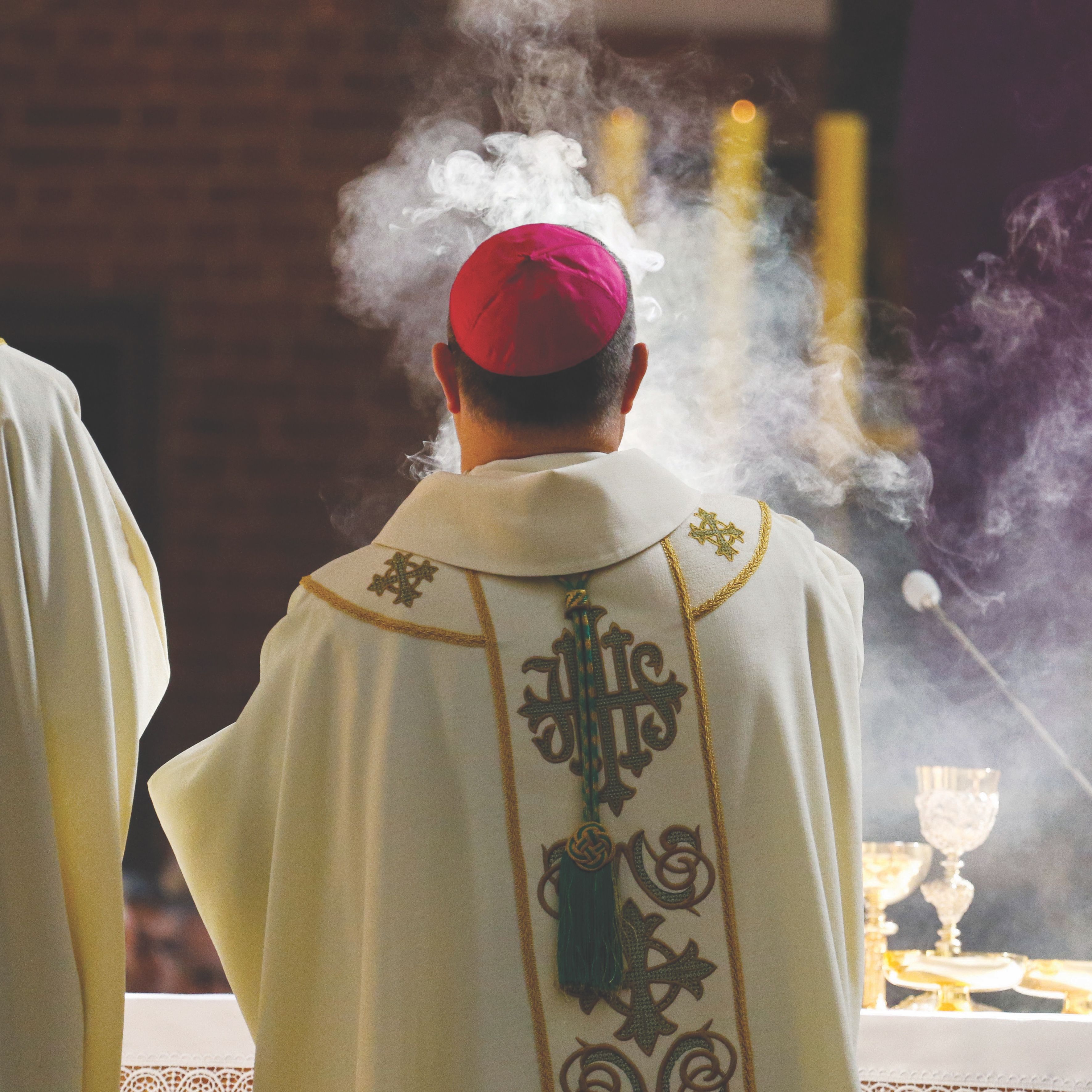 A bishop in ceremonial robes with smoke rising during a religious service.