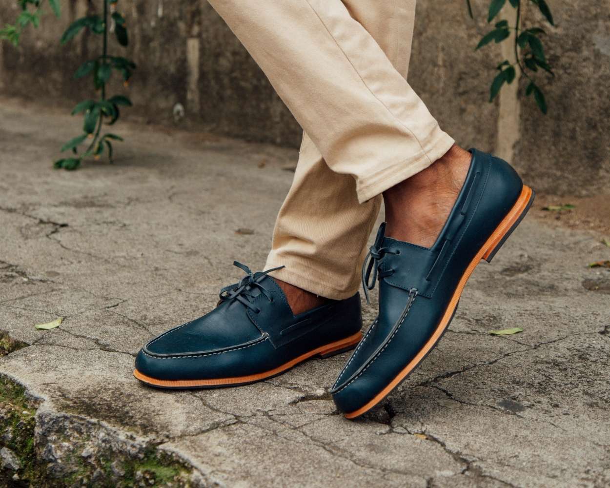 A close-up lifestyle shot of a man's feet walking outdoors, wearing denim blue leather Adelante Nautico boat shoes featuring a distinct light-colored contrast sole and classic laced sides. The man is wearing cream or tan colored chinos, and the background shows a textured concrete or stone walkway with green vines climbing up a rustic concrete wall in the background.